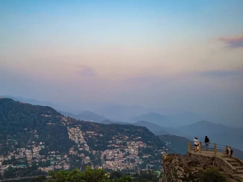 Tiffin Top Nainital panoramic sunset viewpoint over terraced hillside town nestled in misty green mountains hazy purple skies visitors on wooden viewing platform stairs, perfect Uttarakhand tour packages.