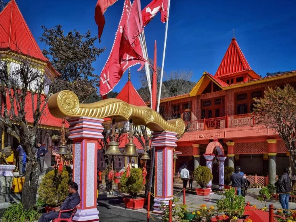 Ornate golden torana gate entrance of Naina Devi Temple Nainital with hanging bells flanked by red-roofed temple buildings, fluttering red flags, pine trees and vibrant flower gardens, perfect spiritual heritage landmark Uttarakhand tour package.