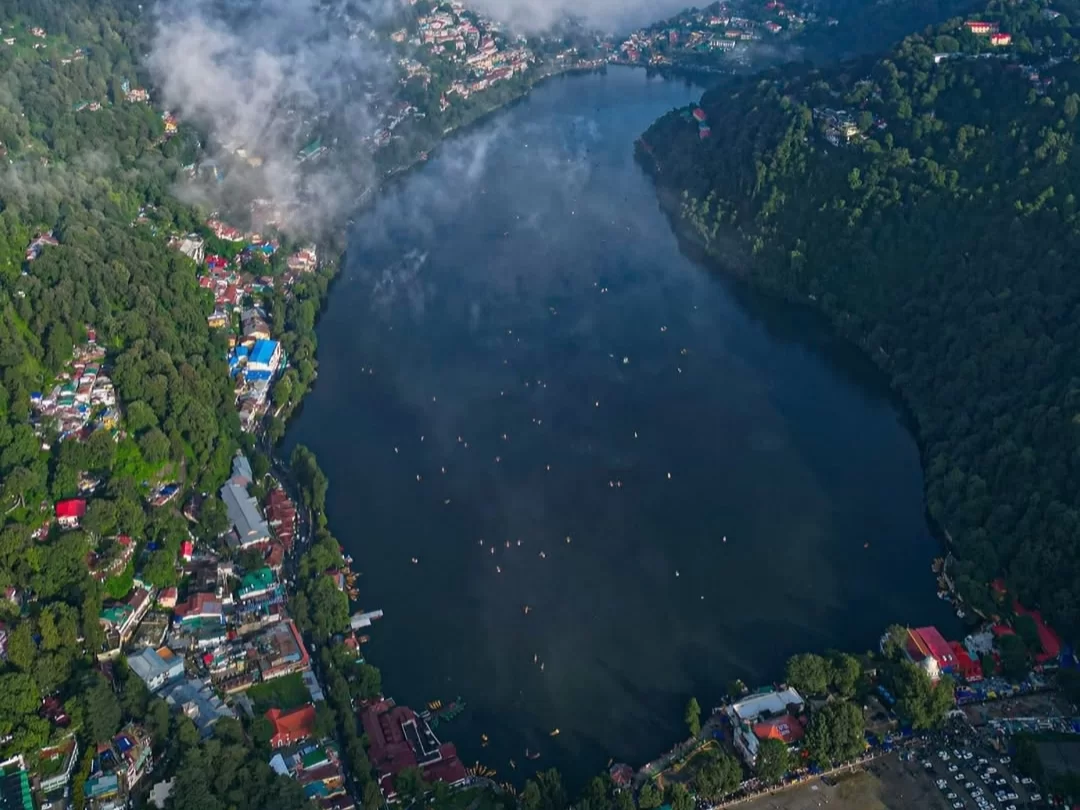 Aerial misty cloudy drone view of Naini Lake Nainital, featuring pear-shaped dark waters, scattered boats, colorful hillside town and forested hills, perfect adventure experience Uttarakhand tour package.