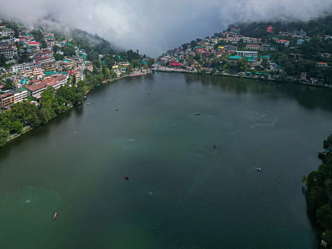 Aerial foggy misty drone view of Naini Lake Nainital, featuring pear-shaped emerald waters, colorful town buildings and forested hills, perfect adventure experience Uttarakhand tour package.