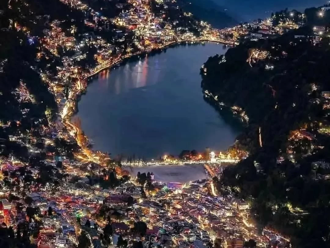 Aerial night view of Naini Lake Nainital, featuring pear-shaped waters reflecting twinkling town lights amid dark hills, perfect romantic experience Uttarakhand tour package.