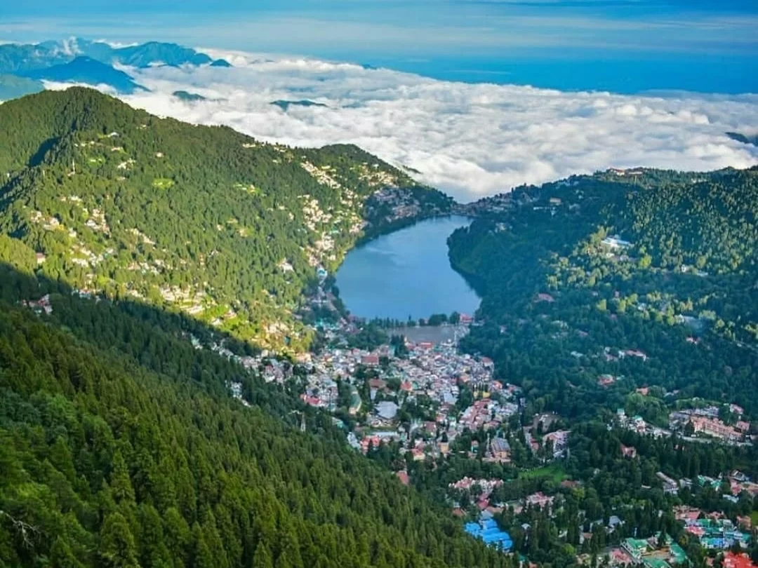 Aerial panoramic of Naini Lake Nainital during cloudy sunny day, featuring pear-shaped turquoise waters amidst sea of clouds and forested hills, perfect romantic experience Uttarakhand tour package.