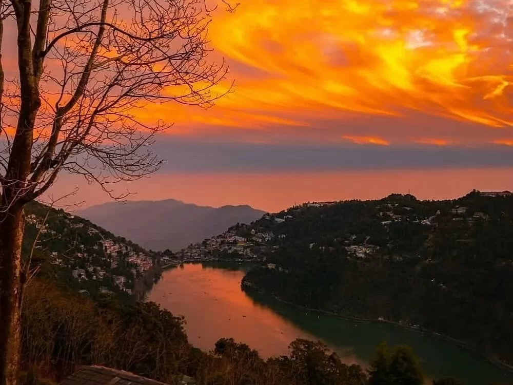 Dramatic orange sunset over Naini Lake Nainital, featuring fiery clouds, pear-shaped waters, town lights and hills, perfect romantic experience Uttarakhand tour package.