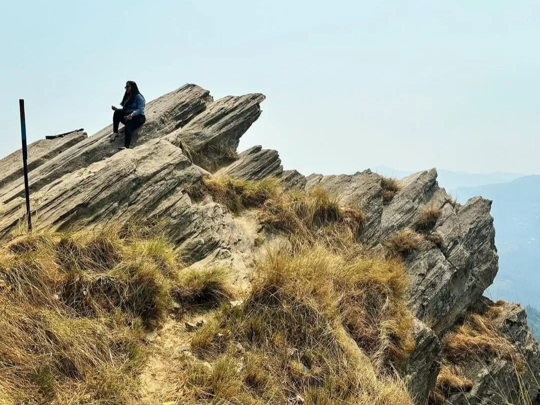 Solo traveler at Chauli Ki Jali during clear day, featuring rocky cliff edge sitting pose grass hills distant mountains pole, perfect adventure trekking experience Uttarakhand Mukteshwar tour packages.