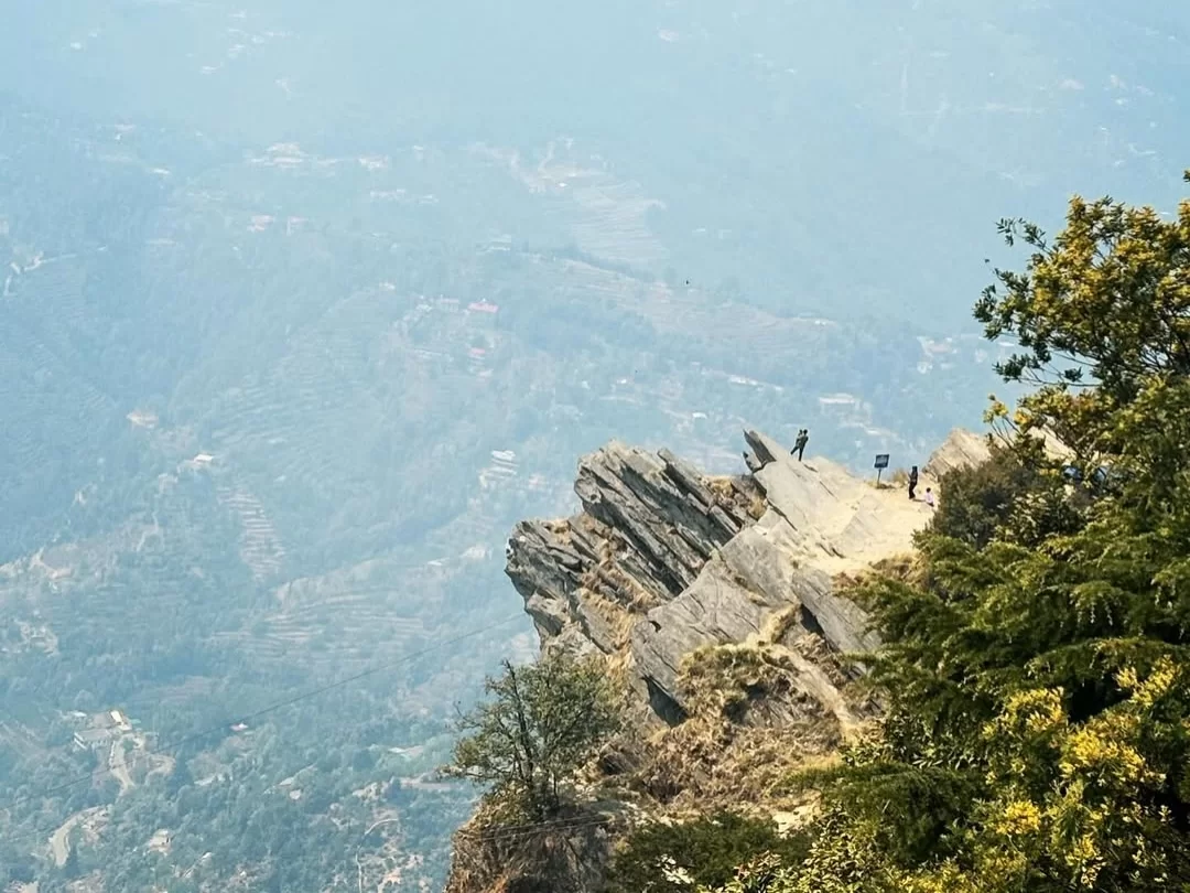Chauli ki Jali viewpoint in Mukteshwar during misty weather, featuring rocky cliff edge, trekkers, Himalayan valleys, perfect adventure spot, Uttarakhand tour packages.