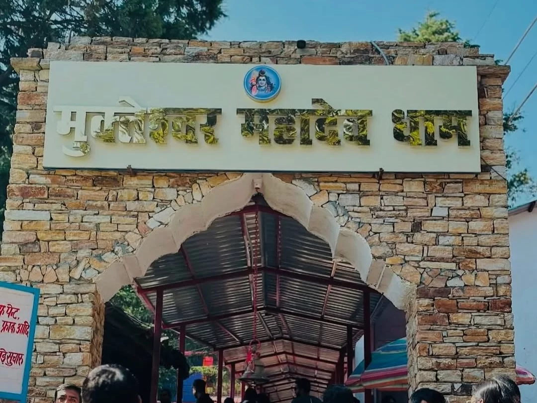 Mukteshwar Mahadev Dham main gate during sunny weather, featuring golden sign Shiva symbol, stone arch, devotees, pine backdrop, perfect spiritual retreat, Uttarakhand tour packages.