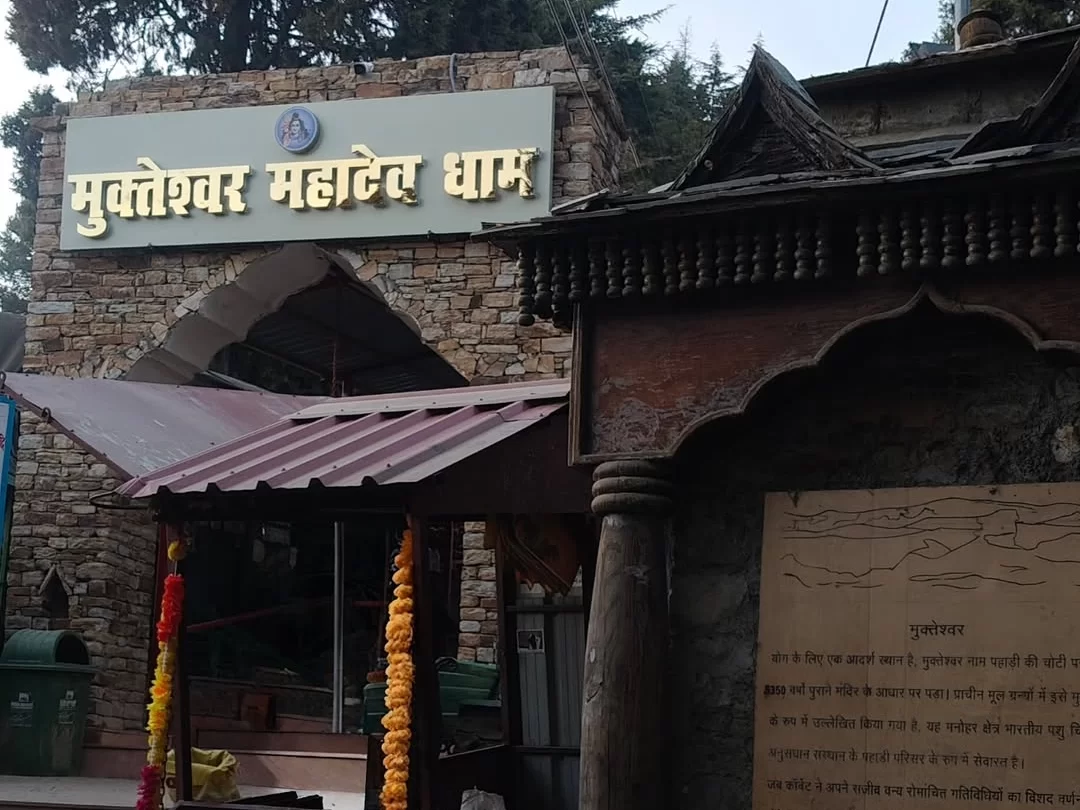 Mukteshwar Mahadev Dham temple gate during misty weather, featuring stone arch, garlands, info board, pine forests, perfect spiritual retreat, Uttarakhand tour packages.