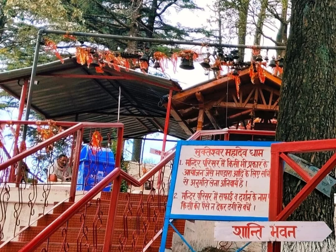 Mukteshwar Dham temple entrance during cloudy weather, featuring red stairs signboard, bells, trees, Shanti Van arrow, perfect spiritual trek, Uttarakhand tour packages.