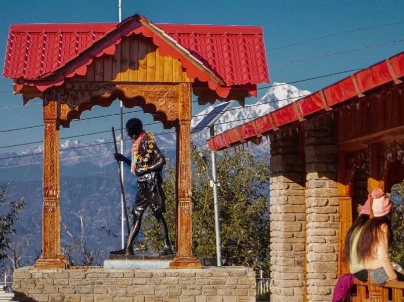Anasakti Ashram Kausani Gandhi statue garlanded staff under red roof wooden pavilion Himalayan peaks background women visitors red roof buildings, perfect Gandhi ashram Himalayan view, Uttarakhand tour packages.