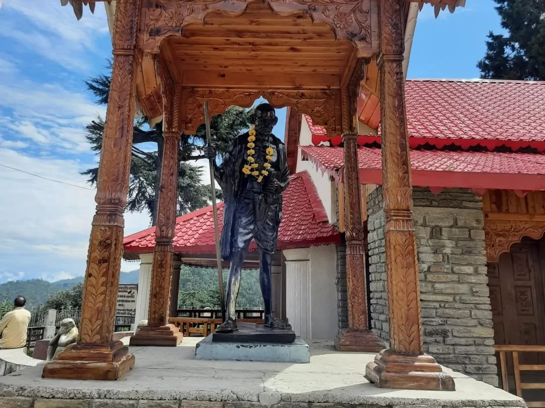 Anasakti Ashram Kausani bronze Gandhi statue garlanded under carved wooden pavilion red roof stone cottage deodar trees blue sky mountains visitors, perfect Gandhi memorial retreat, Uttarakhand tour packages.