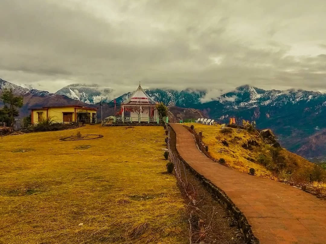 Nanda Devi Temple Ranikhet during cloudy day, featuring white conical temple dome prayer flags buildings yellow grass pathway railing foreground snow-capped Himalayan peaks mist gray sky, perfect spiritual hill station experience Uttarakhand tour packages