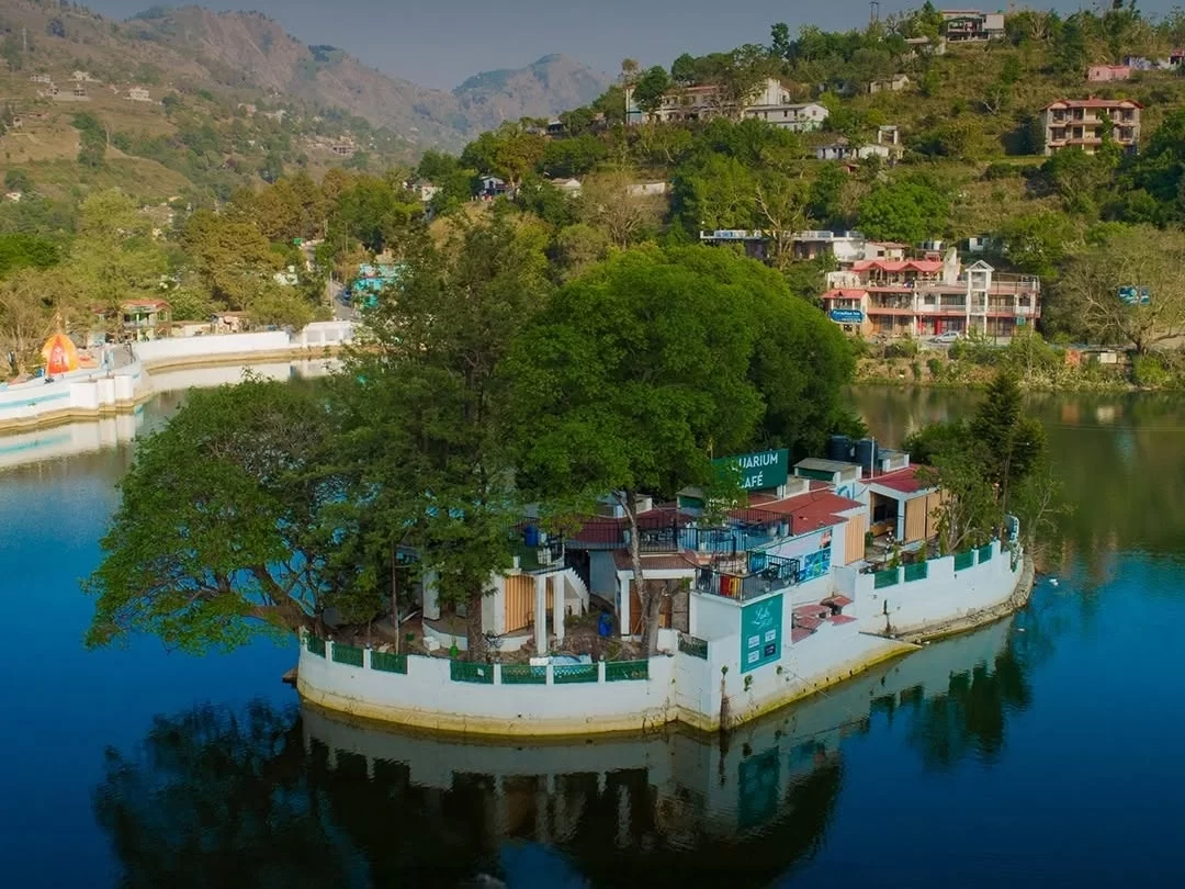 Aquarium Island Cafe on Bhimtal Lake featuring white island structure large trees huts waterfront seating amid hills resorts, perfect Uttarakhand lake adventure tour packages.