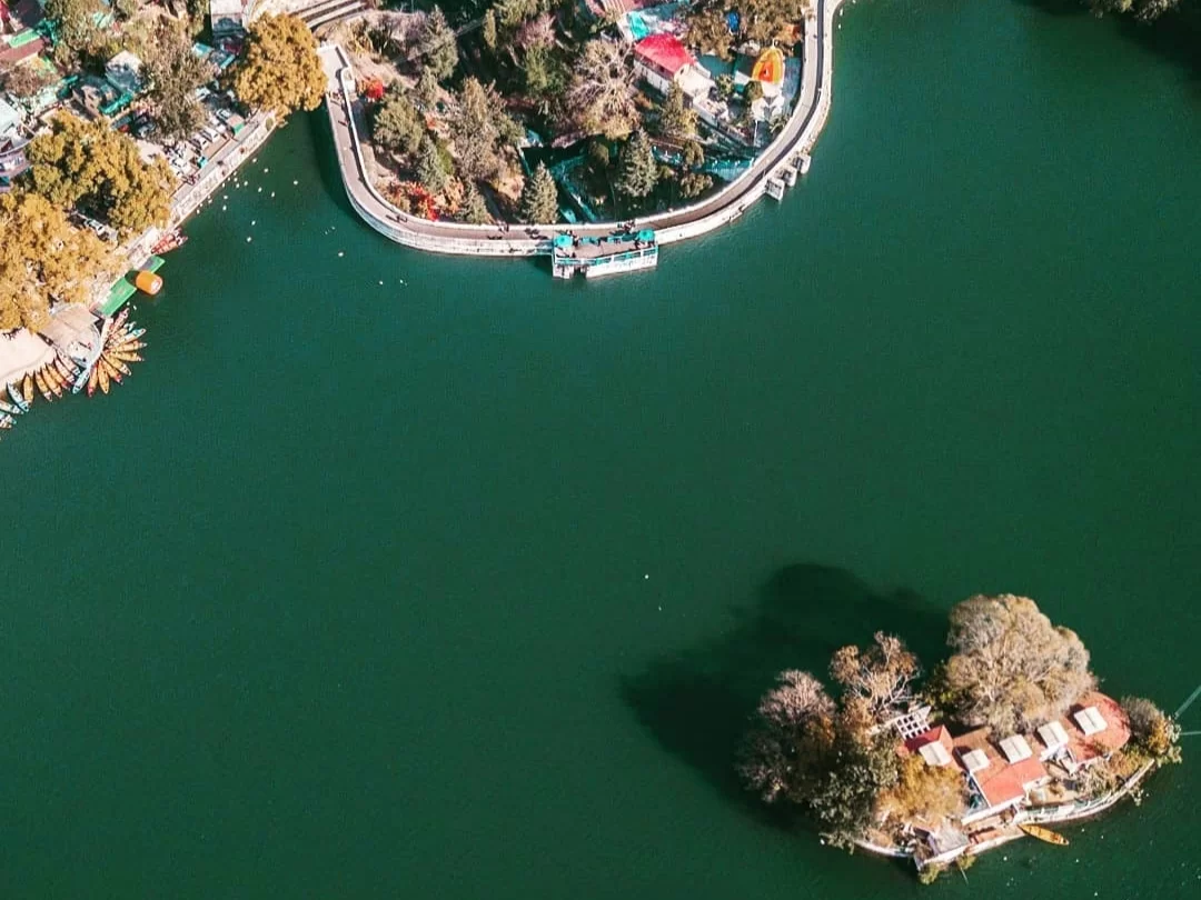 Bhimtal Lake aerial view showing Bhimtal Island, boating jetty with colorful boats & shoreline trees, perfect romantic experience Uttarakhand tour packages. 