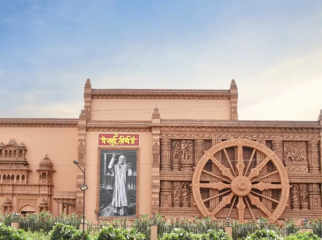 Sai Teerth Devotional Theme Park Chavadi replica during clear day, featuring sandstone structure with large black-and-white Sai Baba image, massive carved Dharma Chakra wheel and yellow Hindi signage amid landscaped greenery, Maharashtra tour package.