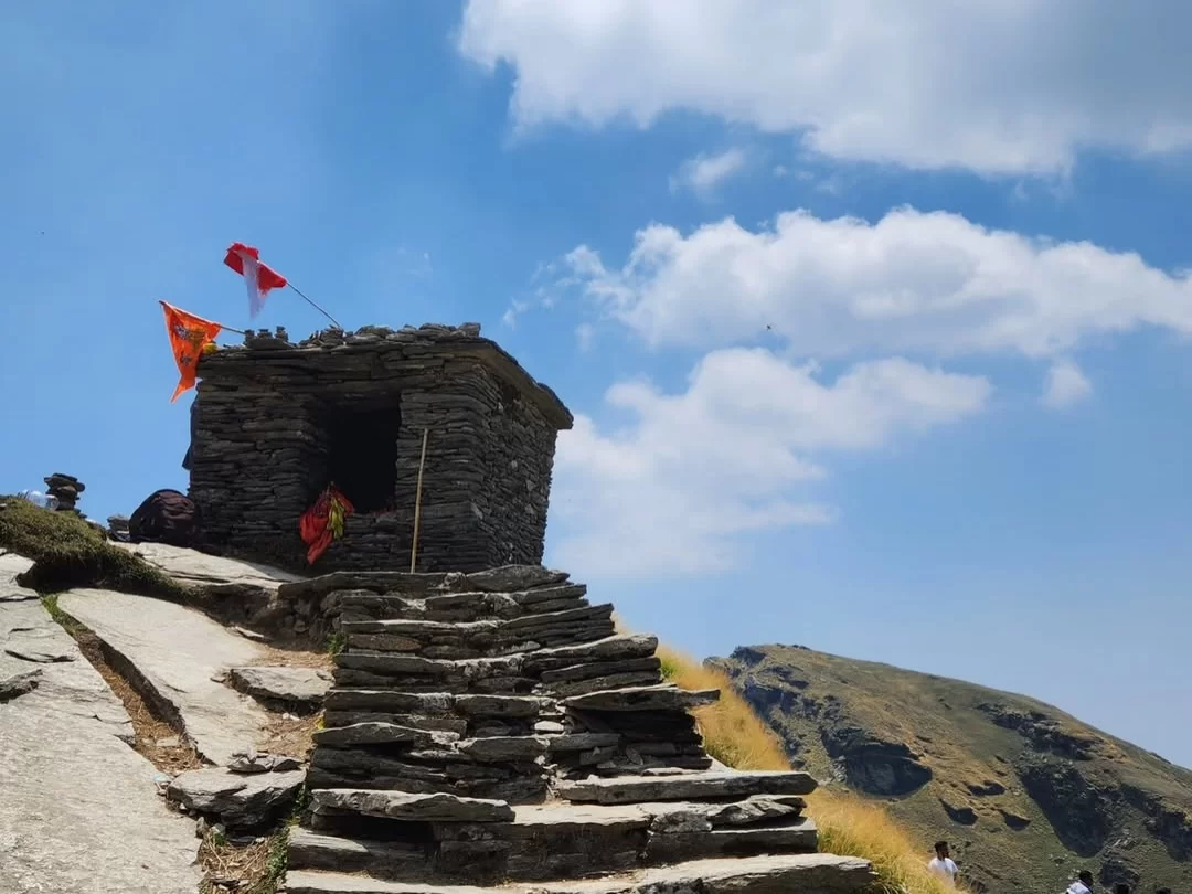 Stone shrine at Tungnath Temple trail during sunny day, featuring saffron flags & hill trek path, perfect adventure experience Uttarakhand tour packages.