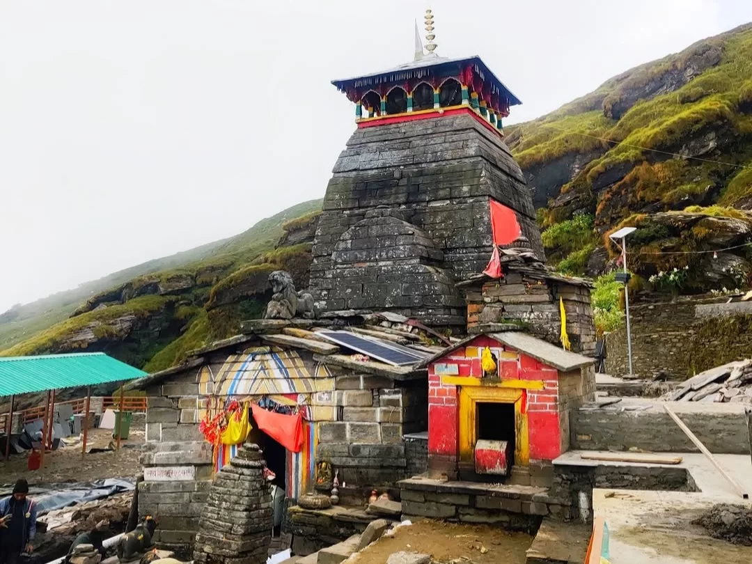 Tungnath Temple shikhar at misty hills during cloudy day, featuring colorful flags & stone facade, perfect adventure experience Uttarakhand tour packages. 