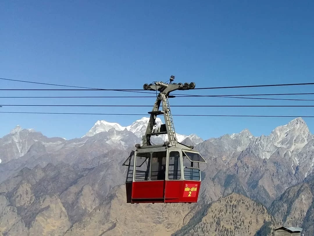 Auli Ropeway red gondola cable car during clear blue sky day, featuring snow peaks Himalayan mountains, perfect adventure experience Uttarakhand tour packages.