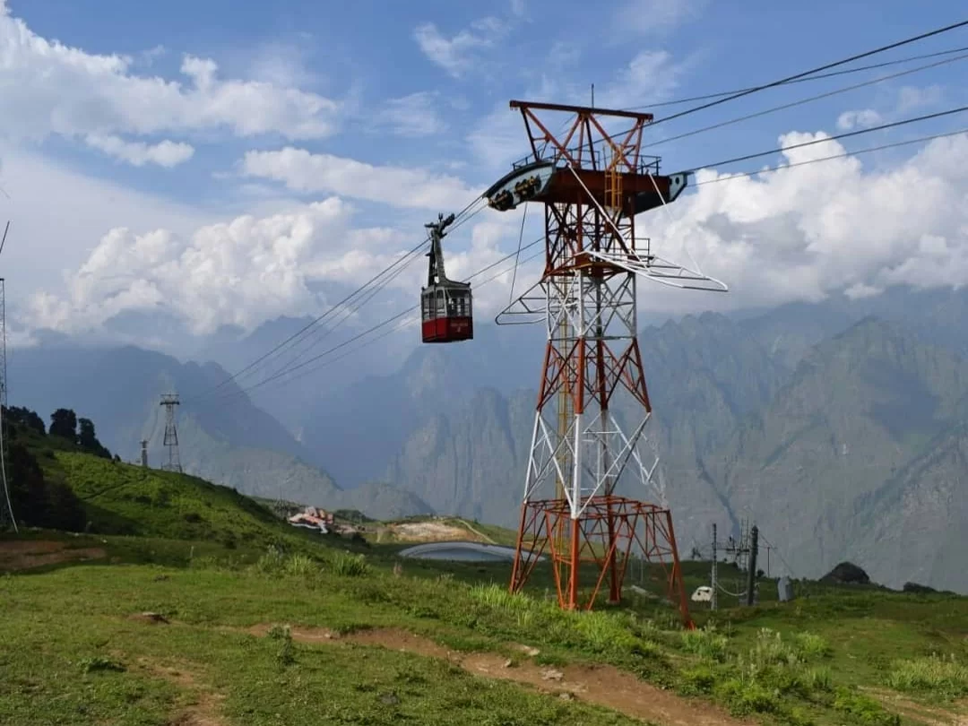 Auli Ropeway red cable car on tower during partly cloudy day, featuring green meadows Himalayan mountains towers, perfect adventure experience Uttarakhand tour packages.