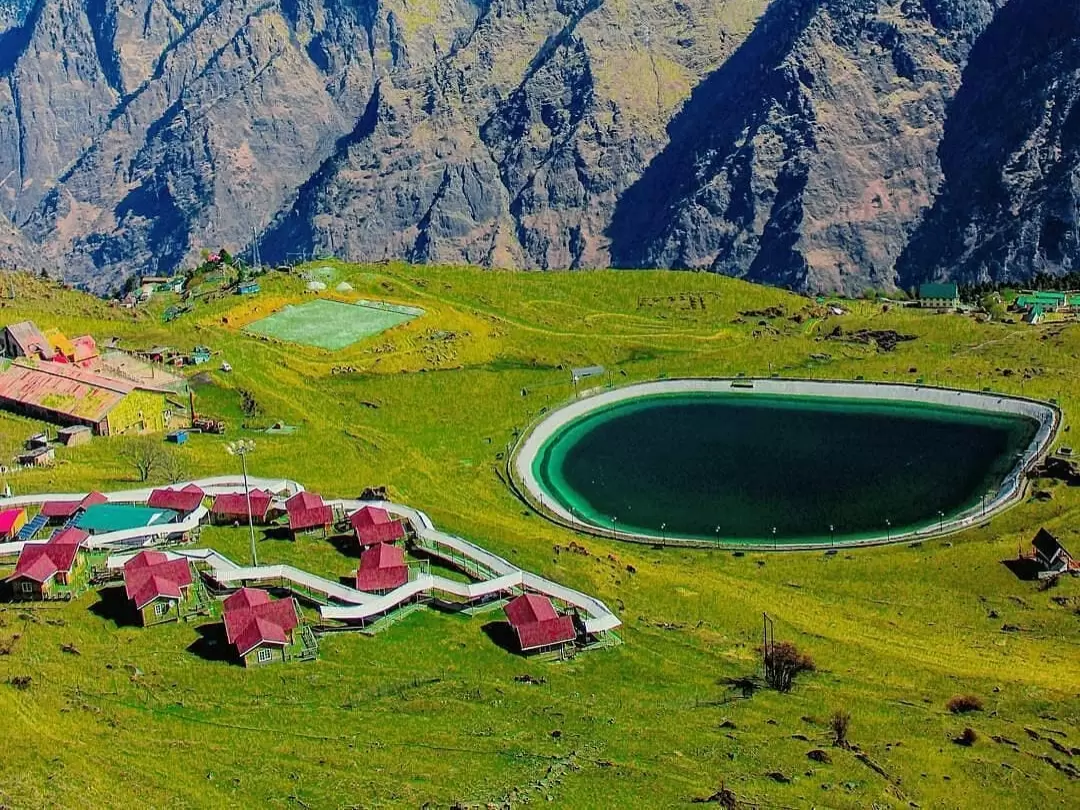 Auli Artificial Lake Uttarakhand vibrant colors green meadows, featuring oval lake red roofed huts paths playing fields Himalayan mountains backdrop, perfect adventure experience Uttarakhand tour packages.