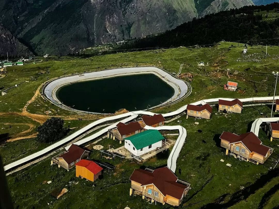 Auli Artificial Lake Uttarakhand aerial view green meadows, featuring oval lake huts chalets paths Himalayan mountains backdrop, perfect adventure experience Uttarakhand tour packages.