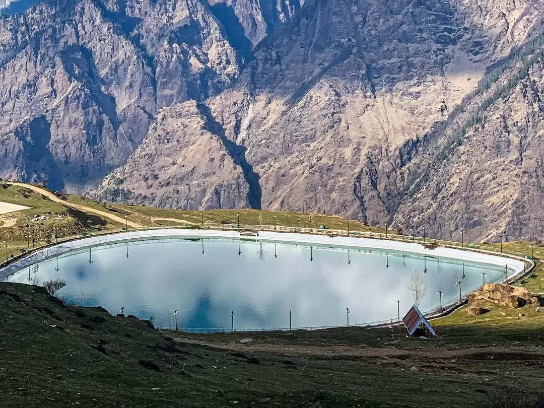 Auli Artificial Lake Uttarakhand during sunny day, featuring blue water snowcapped Himalayan peaks Nanda Devi backdrop walkers railing, perfect adventure experience Uttarakhand tour packages.