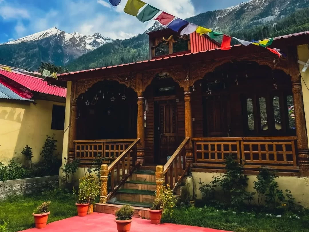 Bagori Village homestay during sunny day Uttarkashi, featuring ornate wooden yellow house prayer flags red pots, snowy mountains blue sky backdrop, perfect Uttarakhand tour packages. 
