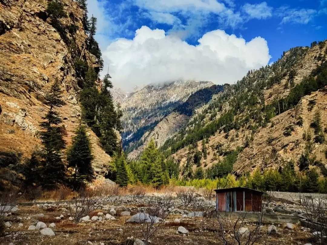 Bagori Village valley landscape during sunny day Uttarkashi, featuring small hut amid pine trees rocky cliffs golden hills snowy distant peaks clouds, perfect Uttarakhand tour packages. 