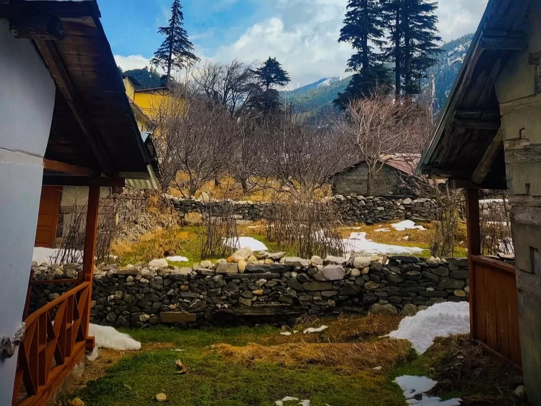 Bagori Village rustic homes during sunny winter day Uttarkashi, featuring stone wall cottages yellow roofs, patchy snow ground pine snowy mountains backdrop, perfect Uttarakhand tour packages.