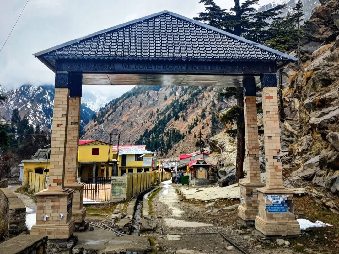 Bagori Village welcome gate during cloudy winter day Uttarkashi, featuring brick archway Welcome to Bagori with colorful houses, snowy rocky mountains pine backdrop, perfect Uttarakhand tour packages.