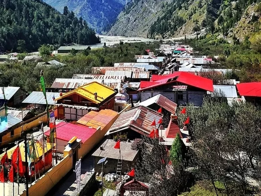 Bagori Village vibrant rooftops during sunny day Uttarkashi, featuring colorful tin-roof houses prayer flags nestled green valley Bhagirathi River snowy mountains backdrop, perfect Uttarakhand tour packages.