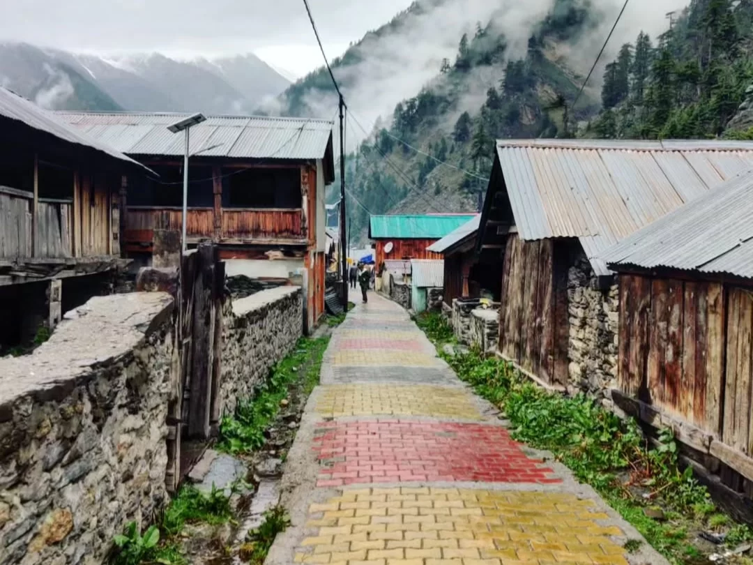 Bagori Village colorful street during misty rainy day Uttarkashi, featuring rustic wooden houses, multi-hued cobbled lane, distant snowy pine hills, perfect cultural adventure Uttarakhand Harshil Valley tour packages.