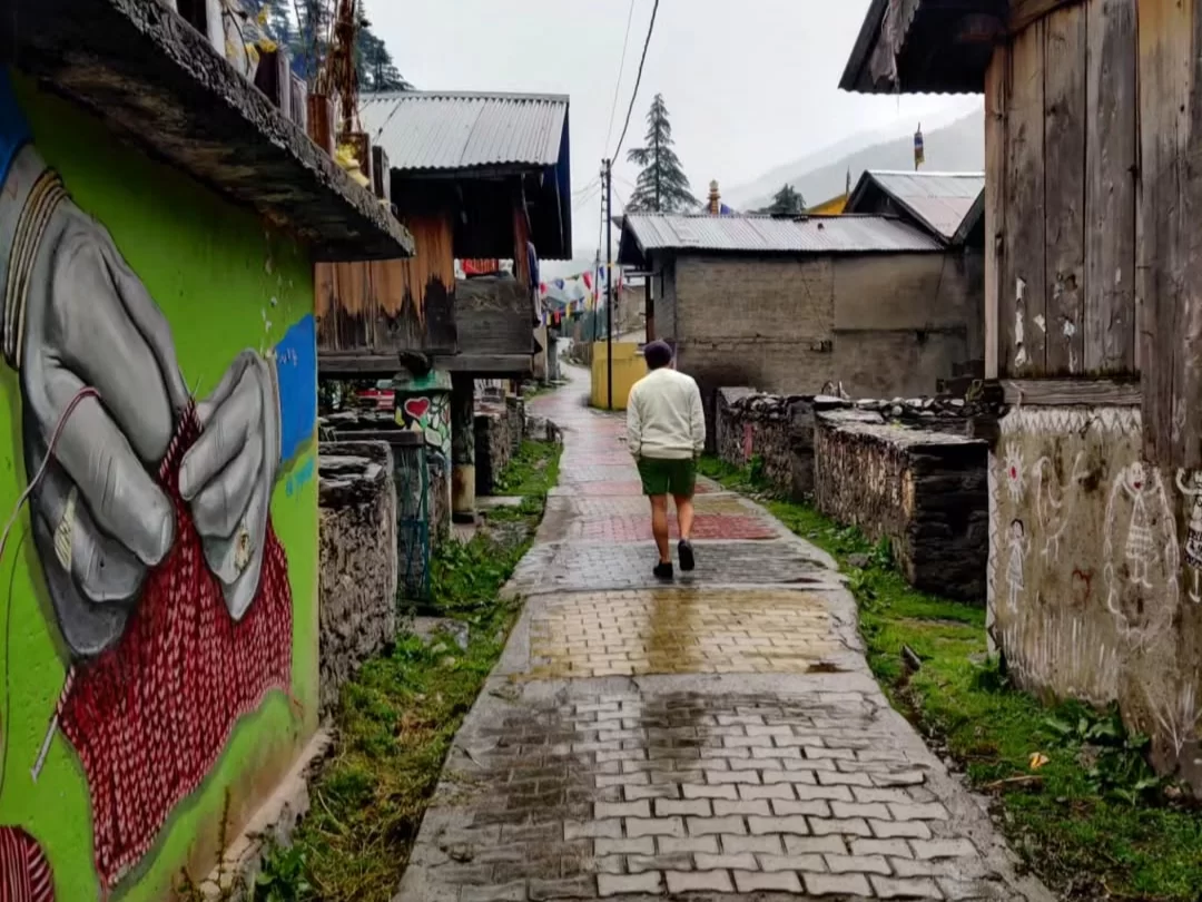 Bagori Village rainy street during misty day Uttarkashi, featuring colorful mural wall art of knitting hands, local man walking wet cobbled lane amid rustic wooden houses pine hills, perfect cultural adventure Uttarakhand Harshil Valley tour packages.
