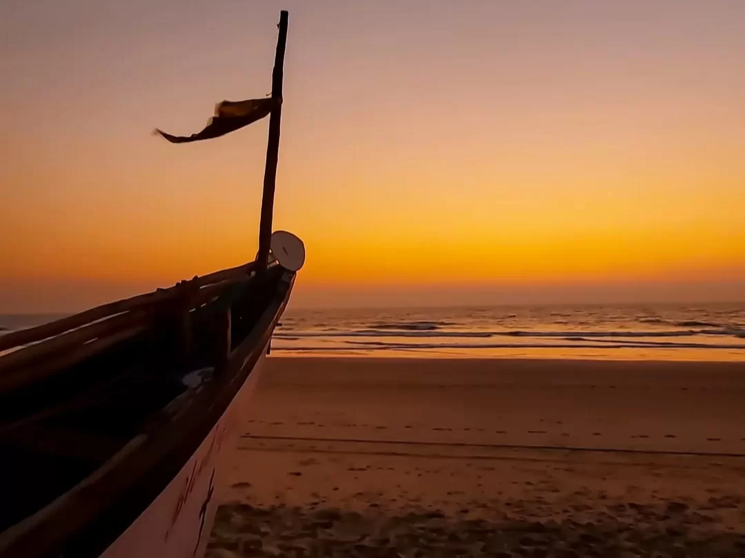 Silhouetted traditional fishing boat flag sunset Betalbatim Beach Goa, featuring orange sky calm waves sandy shore, perfect South Goa beach tour package.