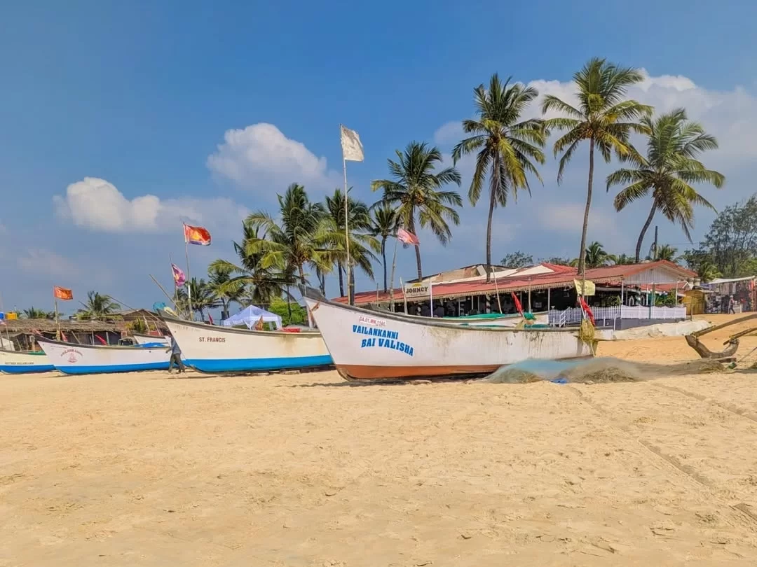 White fishing boat Billauri Bai docked on Benaulim Beach Goa sunny day, featuring flags palm trees beach shacks blue sky, perfect South Goa tour package.