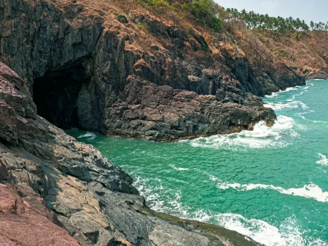 Dramatic sea cave Cabo de Rama Fort Goa cliffside, featuring dark cavern entrance turquoise waves crashing black rocks red earth vegetation, perfect South Goa adventure fort tour package.
