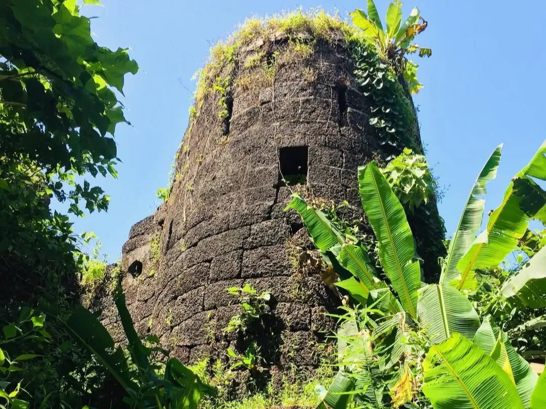 Ancient moss-covered watchtower Cabo de Rama Fort Goa, featuring vine-overgrown stone structure banana leaves clear blue sky tropical forest, perfect South Goa historical fort tour package.