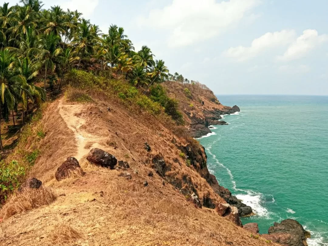Palm-lined dirt trail Cabo de Rama Fort Goa cliff top, featuring red soil path swaying coconuts turquoise Arabian Sea rocky shoreline, perfect South Goa beachside fort trek tour package.
