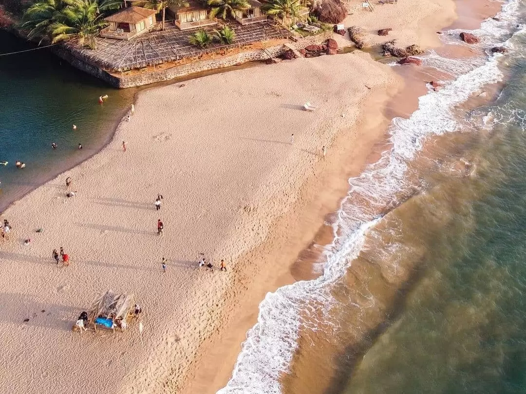 Aerial drone view Cola Beach South Goa sunny afternoon, featuring golden sandy shore turquoise lagoon palm huts people swimming waves, perfect Goa hidden beach tour package. 