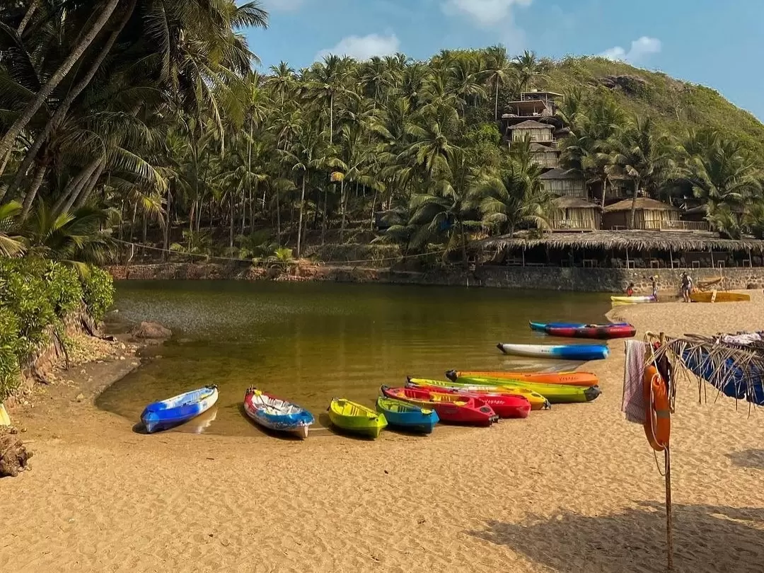 Colorful kayaks lined Cola Beach South Goa partly cloudy, featuring vibrant boats golden sand beach palm-covered hill thatched resorts, perfect Goa water sports beach tour package.