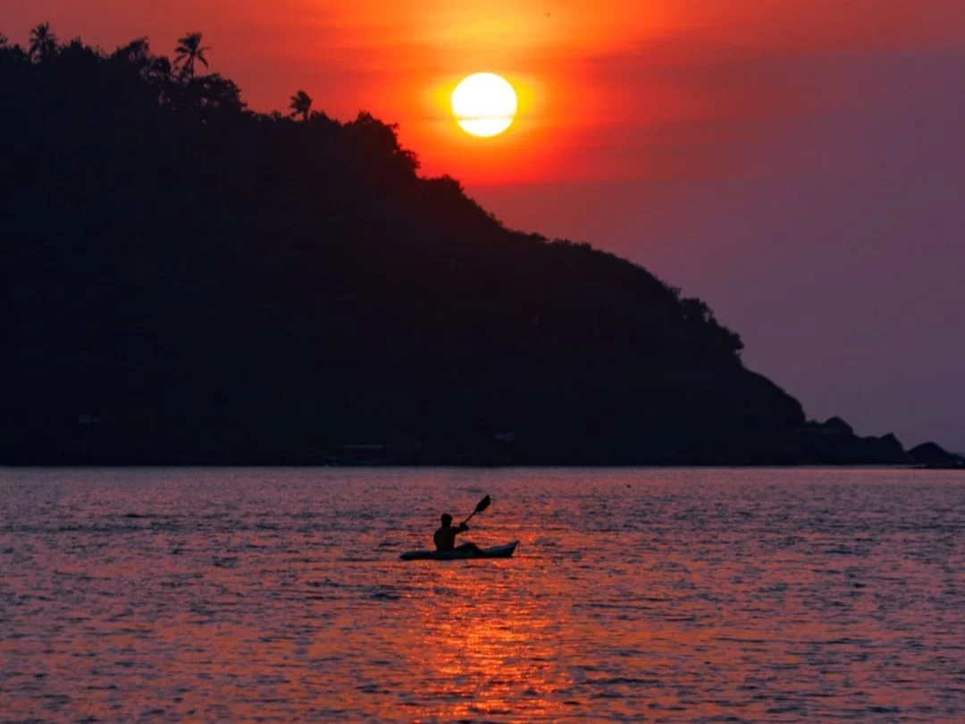 Palolem Beach Goa kayak silhouette fiery sunset Arabian Sea, palm hills golden reflections serene waters, perfect Goa beach paddling dusk adventure package.