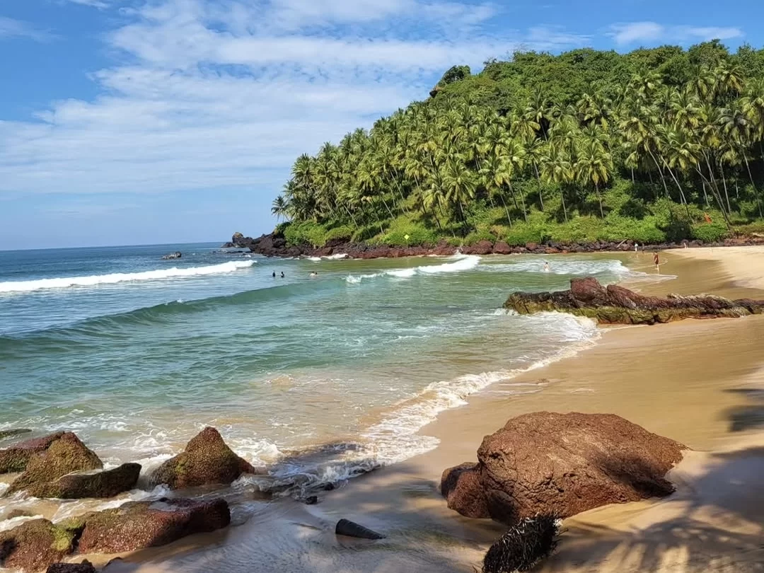 Scenic Caranzalem Beach Goa during partly cloudy skies, featuring palm hill turquoise waves rocky shore swimmers, perfect romantic experience Goa beach tour package. 