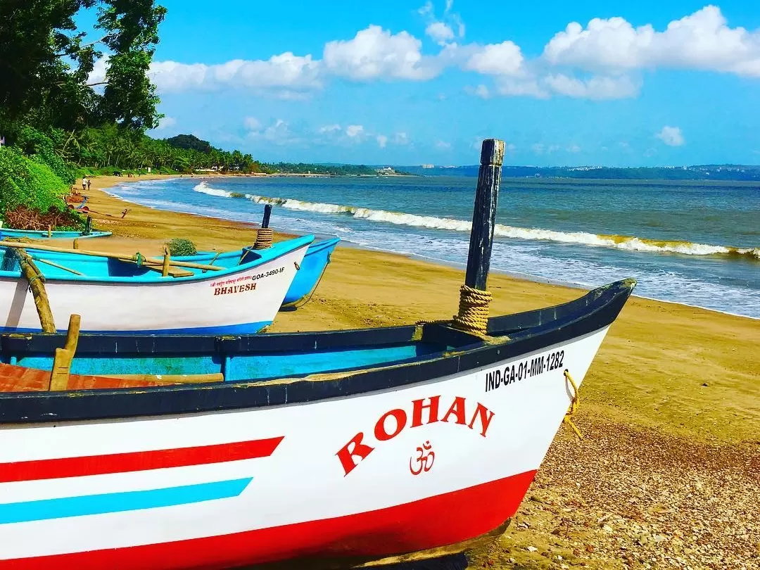 Colorful fishing boats Rohan and others on Bambolim Beach Goa during sunny day, featuring sandy shore, waves and palms, perfect romantic experience Goa beach tour package.