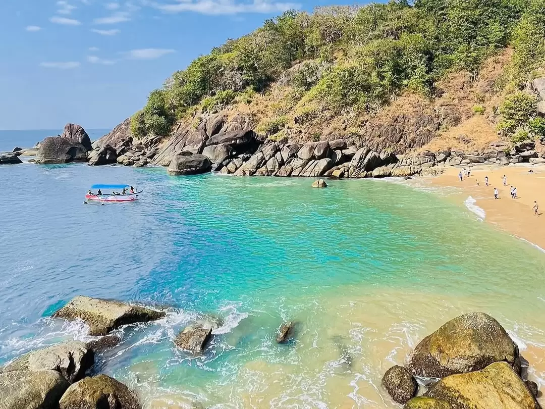 Blue boat and swimmers at Bambolim Beach Goa during sunny day, featuring turquoise waters, rocky cliffs and lush green hills, perfect adventure experience Goa beach tour package. 