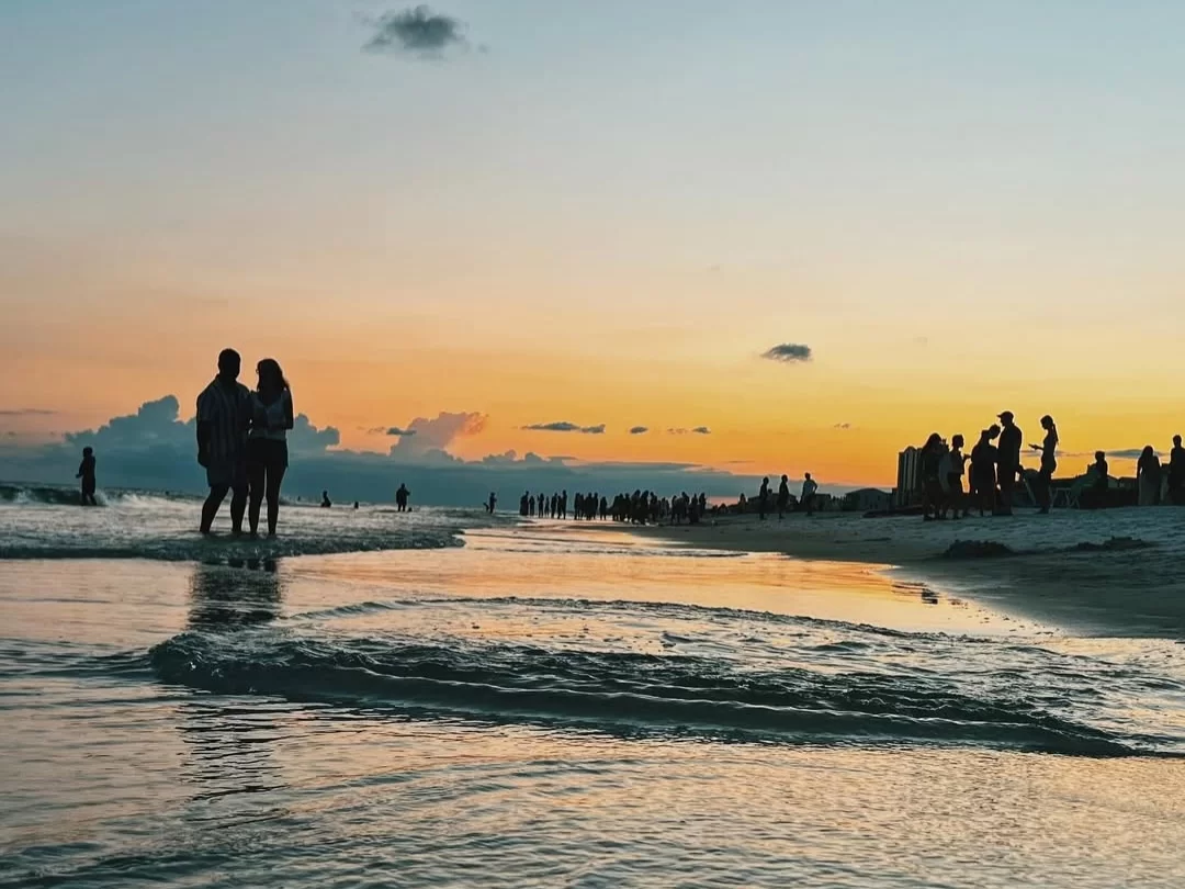 Romantic silhouetted couple holding hands amid beachgoers at sunset Miramar Beach Goa, featuring orange sky gentle waves sandy shore crowds, perfect beach experience Goa romantic sunset beach walk package. 