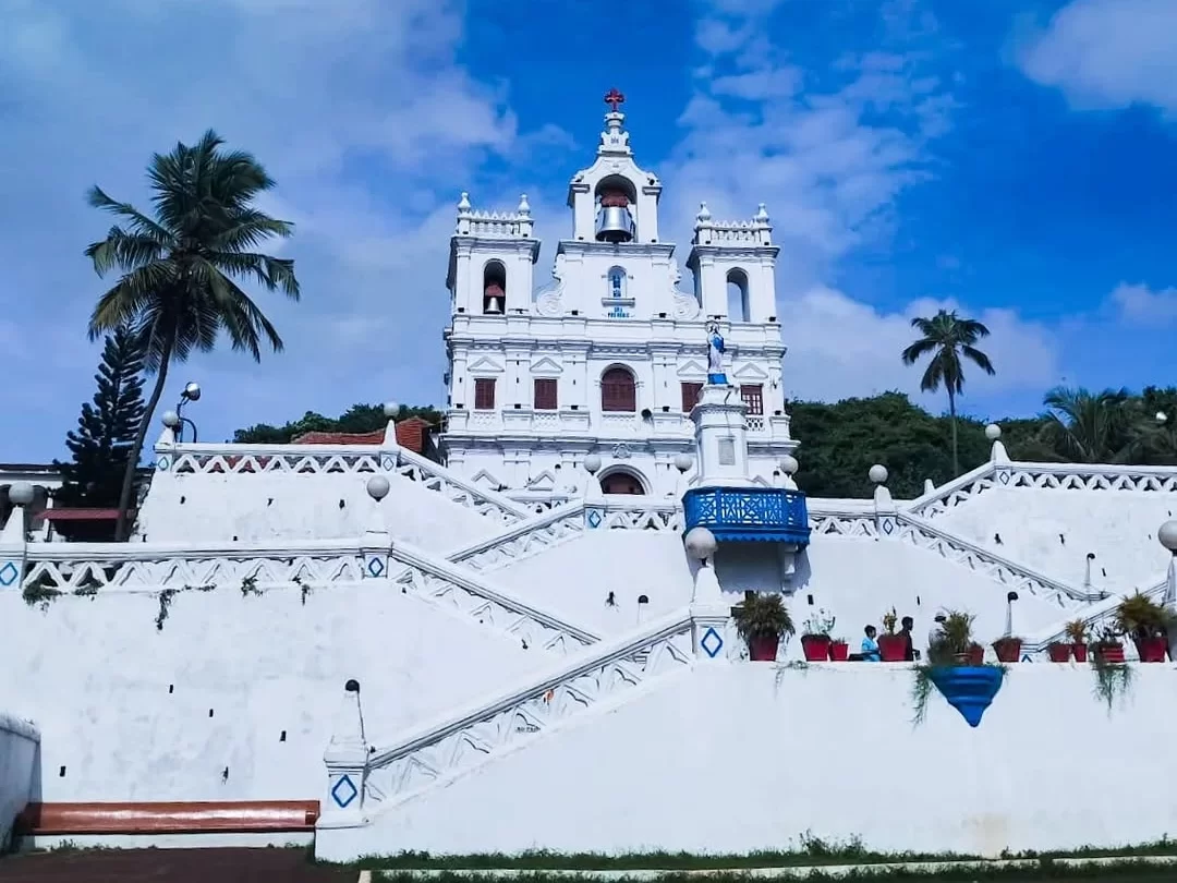 Bright Our Lady of Immaculate Conception Church Panjim Goa under partly cloudy blue sky with visitors potted plants on sweeping white staircase palm trees blue balustrade lamps greenery accents, perfect heritage experience Panjim Goa church feast Immacula