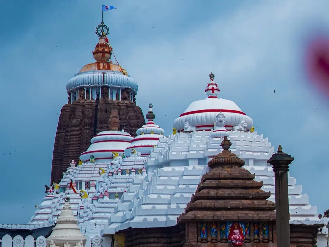 Shree Jagannath Temple towers at Puri during partly cloudy day, featuring Neelachakra flag, white-red domes, ancient spires, perfect spiritual experience with Odisha Puri tour package.