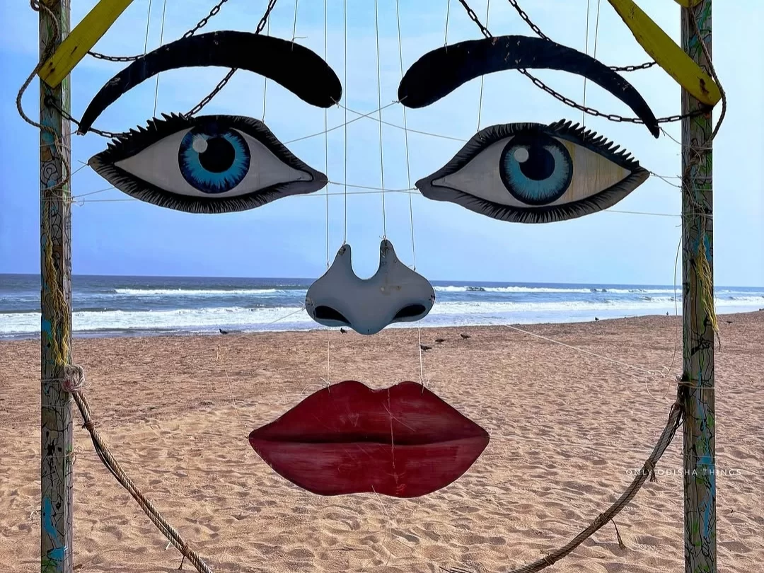 Puri Beach Odisha giant colorful face sculpture with oversized blue eyes black eyebrows red lips yellow chain frame against Bay Bengal waves golden sands, iconic Sudarsan Pattnaik-style beach art installation perfect Jagannath Puri India tour package. 