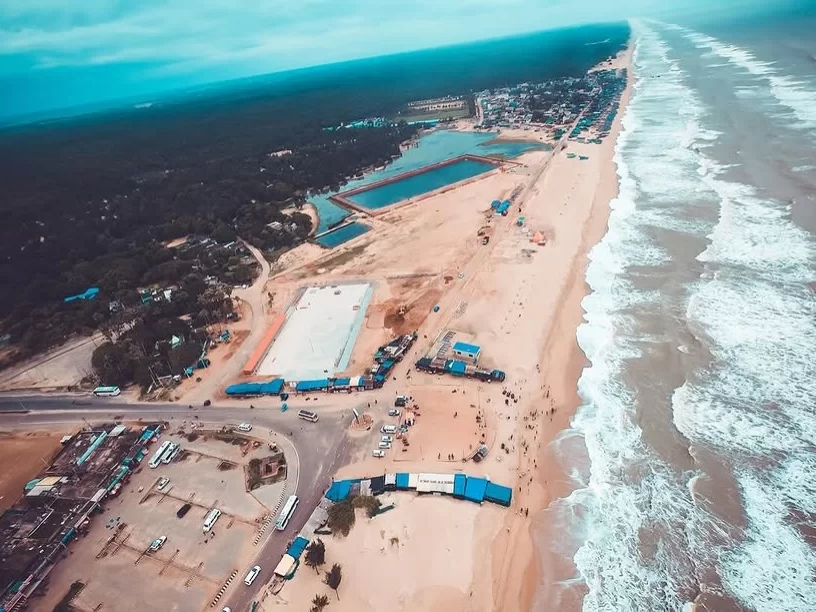 Chandrabhaga Beach Odisha aerial drone view featuring blue water tanks sandy beach road palm groves fishing village ocean waves cloudy sky near Konark Sun Temple, perfect beach experience for Konark Chandrabhaga tour package.
