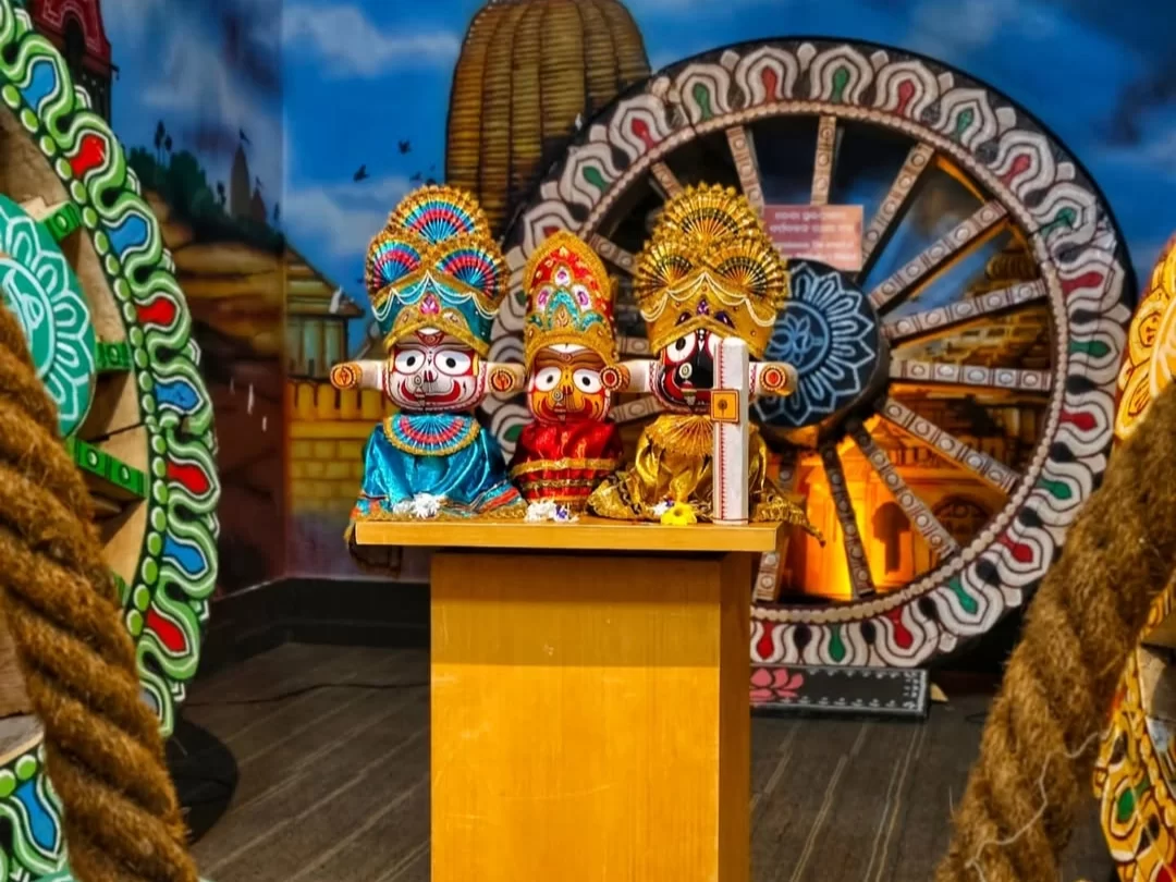 Odisha State Museum Bhubaneswar Jagannath Balabhadra Subhadra idols Ratha Jatra chariot wheel backdrop thatched huts, perfect Odisha tribal heritage culture tour package.