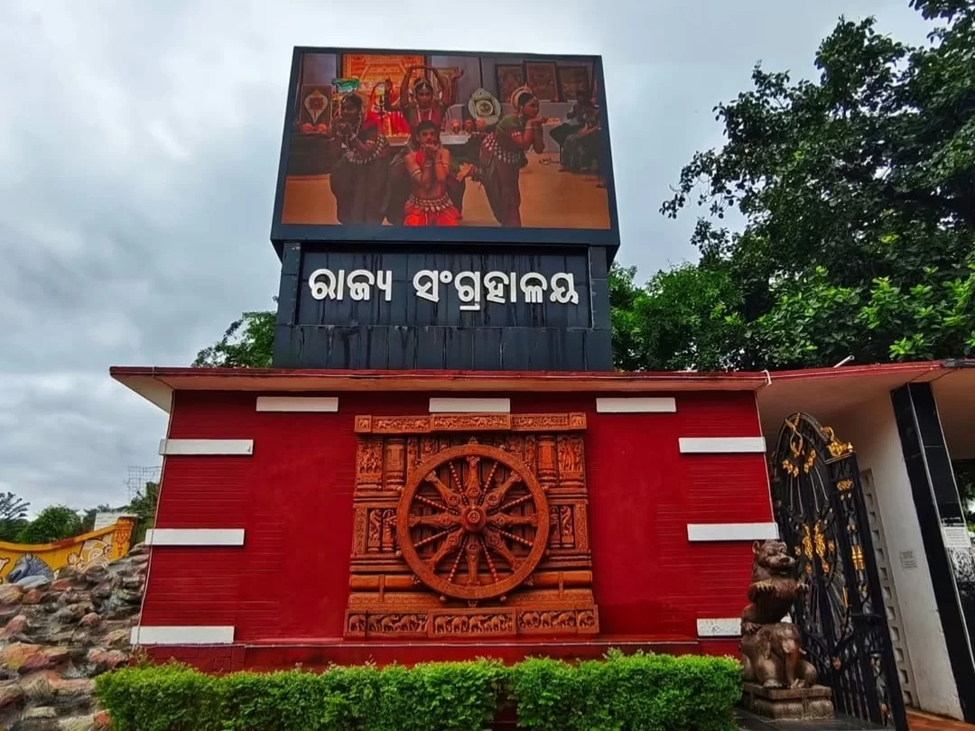 Odisha State Museum Bhubaneswar grand red entrance gate Odisha script LED tribal dancers chariot wheel cloudy sky, perfect Odisha heritage culture tour package. 